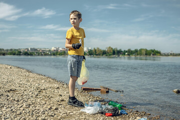 A child boy volunteer in gloves tidying up rubbish plastic garbage from the riverbank. Children take care of nature