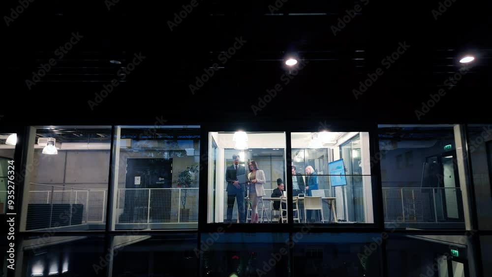 Poster Businessman and a Female Executive Manager Standing Next to a Window in Office, Using a Laptop Computer. Team of Specialist Having a Meeting, Discussing a Report. View From Outside the Window at Night