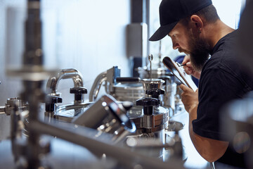Bearded brewery technologist, focused man inspecting stainless steel brewing equipment, taking notes on clipboard. Craftsmanship of beer production. Beer, brewery, manufacture, quality control concept