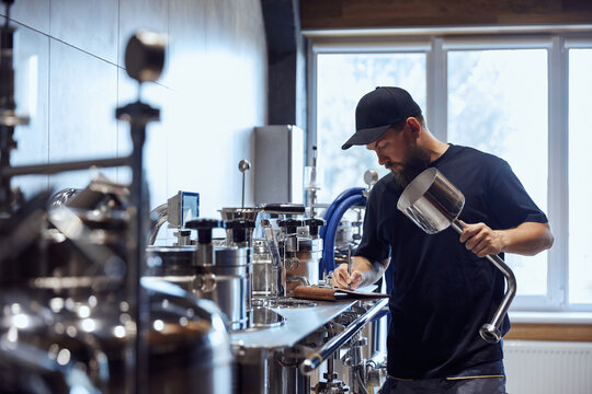 Standing in brewery, technologist taking notes during checking brewing process and equipment. Science and craft in beer production. Concept of beer, brewery, manufacture, quality control - Powered by Adobe