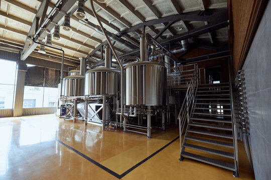 Large, modern brewery with several stainless steel tanks, connected by a network of pipes and valves, with staircase leading to platform above. Concept of beer, brewery, manufacture, quality control