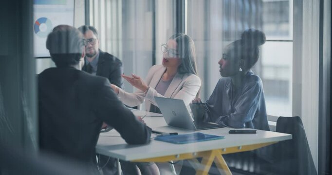 Young Businessmen and Businesswomen Having Negotiations in the Office in a Glass Room. Female Entrepreneur Sitting Behind a Meeting Table, Discussing Statistics and Analytics Data with the Team