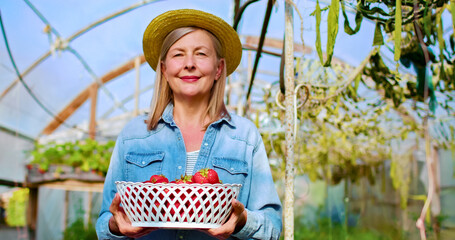Close up portrait of old Caucasian woman wearing straw hat holds basket full of freshly picked strawberries in hands. Happy beautiful female farmer smiles enjoying summer harvest.