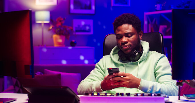 Close up portrait of young African male with headphones on neck holds phone in hands chatting with friend. Handsome bearded face man sits at table with different gadgets in LED lighted room.