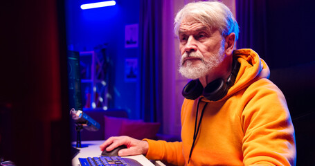 Close up portrait of old Caucasian male with headphones on neck looks at computer screen in LED lighted room. Handsome bearded face man works at home using gadgets.
