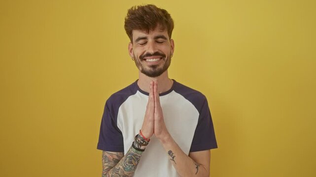 Confident young hispanic man standing over a yellow isolated background, praying with hands together, asking for forgiveness
