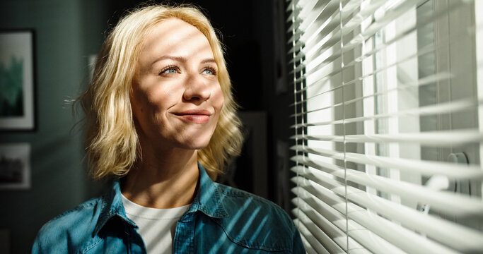 Close up portrait of beautiful young Caucasian female looking out window with smile on face. Sun's rays fall on woman's face with warm.