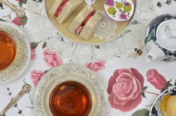 Still life with tea and sweets: traditional english tea party at five o'clock with triangular sandwiches and cupcake on plateau. Top view. Tablecloth with roses.