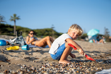 Young Boy Playing with Shovel on Pebble Beach While Mother Relaxes in the Background