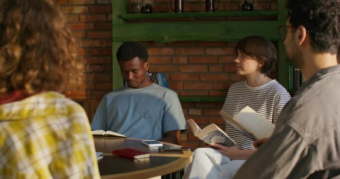 Young people of different nationalities read the Bible while sitting at a table indoors. They are holding books in their hands. Daylight