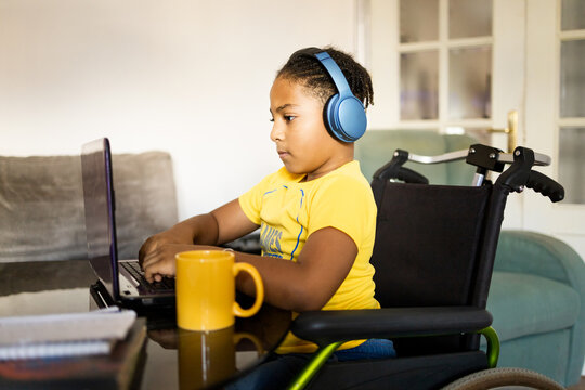 A dark-skinned boy between 8 and 9 years old sitting in a wheelchair is using a laptop in his living room. The disabled African child is studying with a computer.Children in wheelchair concept.