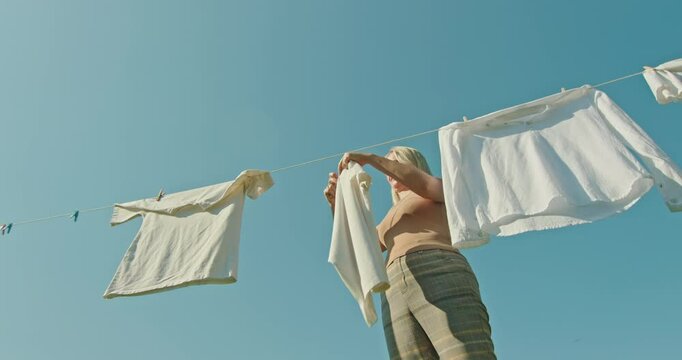 Middle-aged woman hangs wet white clothes on a clothesline for drying, viewed from below against a blue sky.