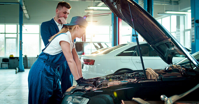 Portrait of two employees wearing specialized clothes work at service station. Young people examine car under hood to find breakdown. - Powered by Adobe