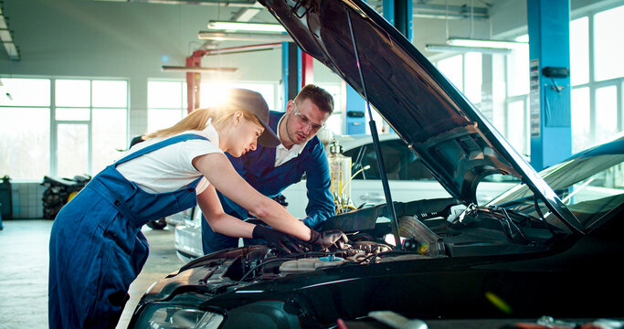 Portrait of two employees wearing specialized clothes work at service station. Young Caucasian people repair car helping each other.