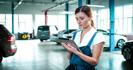 Close up portrait of young Caucasian female wearing specialized clothes stands in the middle of service center with tablet in hands. Beautiful woman makes report of working day.