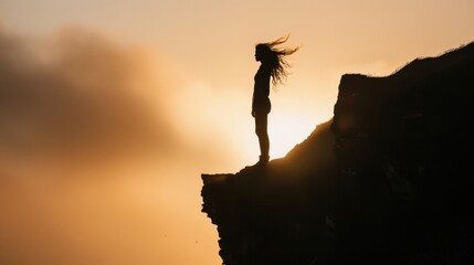 Naklejka premium A person standing on a cliff edge, backlit by the setting sun, with the wind blowing dramatically through their hair