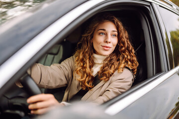 A young woman with curly hair smiling while driving a car in a city during daytime on a clear sunny day