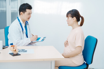Asian female doctor listens to belly of pregnant mother during a prenatal exam in clinic. doctor...