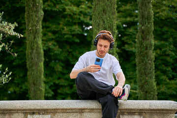 A man is sitting on a stone wall with his cell phone in his hand. He is wearing headphones and he is listening to music