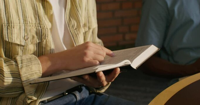A man holds Bibles in his hands, sitting at a table indoors, close-up of his hands, unrecognizable person. Daylight