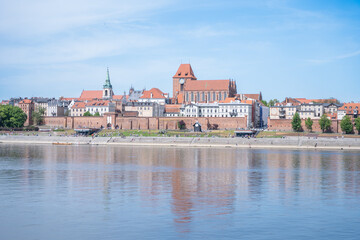 A panoramic view of the old town of Toruń, Poland, on a sunny day, showcasing its medieval architecture, red brick buildings, and the picturesque Vistula River. 