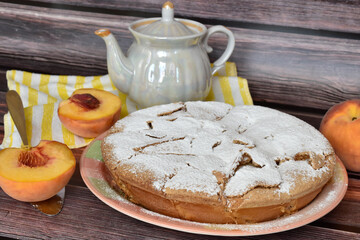 Fresh baked homemade charlotte pie sprinkled with powdered sugar. Peach half on metal cake shovel, juicy peach fruit, pearlescent teapot, striped yellow and white napkin on wooden background. 
