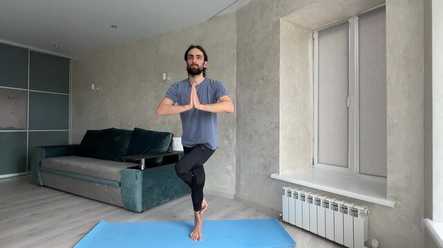 Yogi man maintains balance while practicing the eagles pose in a cozy living room setting, promoting fitness and serenity. Garudasana