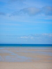 A sandy beach at low tide, with a blue ocean and a blue sky with clouds. A small boat is on the ocean.