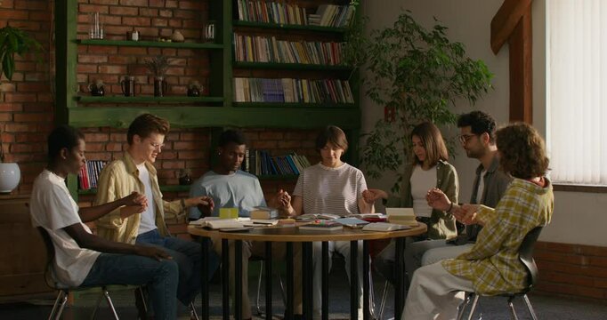 Young people of different nationalities pray holding hands, sitting at a table indoors. Daylight