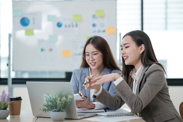 Two women are sitting at a table with a laptop and a whiteboard