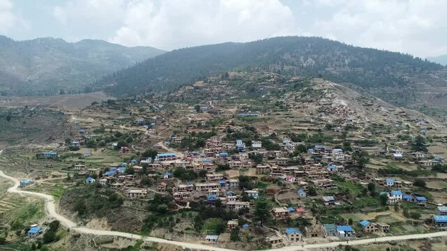 "Aerial view of a traditional village in Jumla, Nepal, nestled in the hills. The village features houses with blue roofs scattered across terraced fields, surrounded by rugged terrain and lush forests