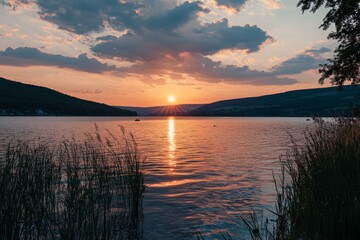 Obraz premium A stunning sunset over a calm lake with the sun's reflection on the water, surrounded by reeds. The sky is painted with shades of orange and pink, creating a serene atmosphere.