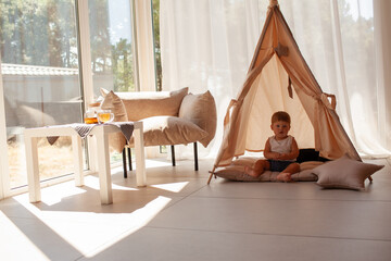 Small child under one year old plays in the room against the backdrop of wigwam in beige shades. The infant baby is happy and dressed  bodysuit. The cozy home environment © Вероника Зеленина