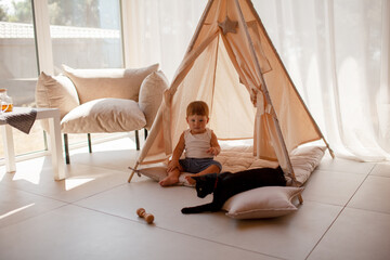 Small child under one year old plays with cat in the room against the backdrop of wigwam in beige shades. The infant baby is happy and dressed  bodysuit. The cozy home environment © Вероника Зеленина