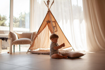 Small child under one year old plays in the room against the backdrop of wigwam in beige shades. The infant baby is happy and dressed  bodysuit. The cozy home environment © Вероника Зеленина