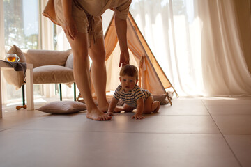Small child under one year old plays in the room against the backdrop of wigwam in beige shades. The infant baby is happy and dressed  bodysuit. The cozy home environment © Вероника Зеленина
