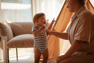 Small child under one year old plays in the room with mom against the backdrop of wigwam in beige shades. The infant baby is happy and dressed  bodysuit. The cozy home environment © Вероника Зеленина