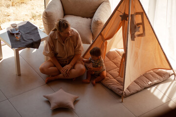 Small child under one year old plays in the room with mom against the backdrop of wigwam in beige shades. The infant baby is happy and dressed  bodysuit. The cozy home environment © Вероника Зеленина