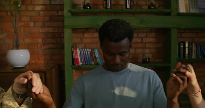 Young people of different nationalities pray holding hands, sitting at a table indoors. Daylight
