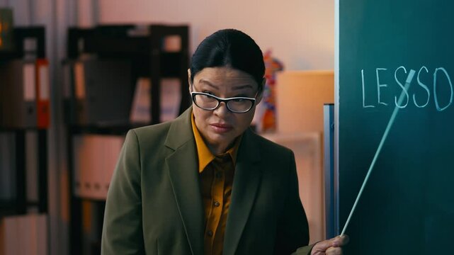 Comically angry teacher in her 50s pointing at chalkboard during lecture, comedy