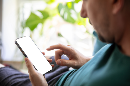 Man holding smartphone with blank screen while relaxing on sofa at home. Bright greenery background. Ideal for UI/UX design templates and app mockups