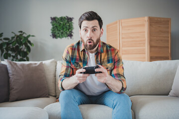 Photo of handsome young guy sit couch amazed play video game dressed plaid shirt spend pastime spacious house indoors room