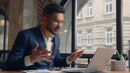 Fototapeta premium Stressed mad furious angry Arabian Indian man businessman male guy business leader boss talking video call online conference chat laptop at office shout scream yell problem quarrel conflict at office