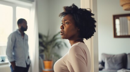 A woman stands in a softly lit room, looking away thoughtfully, as a man stands in the background, suggesting an emotion-laden moment.