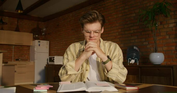 A man is studying the Bible while sitting at a table indoors. He prays with his hands folded in front of him. Daylight