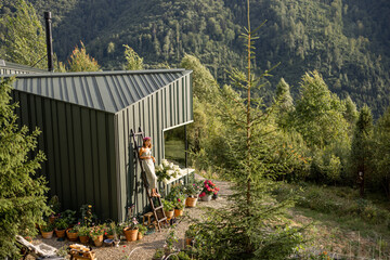 A woman stands on a ladder beside a modern eco-house surrounded by mountains and a blooming garden. The morning light adds warmth, highlighting the beauty of nature and the tranquility of rural life