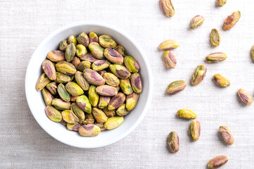 Pistachios kernels, roasted and salted nuts in a white bowl on linen fabric. Shelled fruits of Pistacia vera, crunchy toasted and salted. Ready-to-eat snack food. Close-up, from above, food photo.