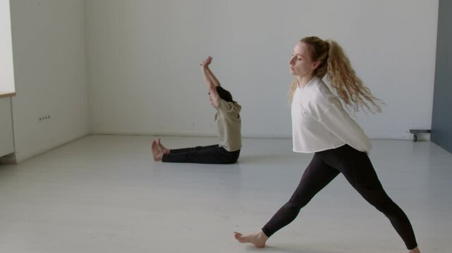 Graceful female dancer in leggings and sweatshirt doing wide legged standing bend in studio while male partner practicing forward fold and lying down hamstring stretch in background