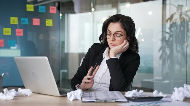 Bored female office worker sitting at desk with laptop and crumpled paper, looking tired. Woman disengaged in office environment. Workplace frustration and lack of motivation in a professional setting