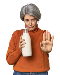 Caucasian mid-age female holding milk bottle standing with outstretched hand showing stop sign,...